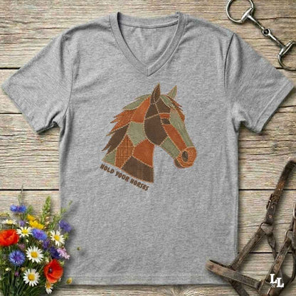 A grey V-neck t-shirt laid flat on a rustic wooden surface, featuring a stylized patchwork horse head graphic with the text 'HOLD YOUR HORSES' underneath. The shirt is styled with equestrian tack, a small bouquet of wildflowers, and a metal horse bit.
