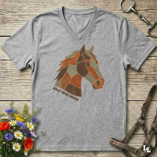 A grey V-neck t-shirt laid flat on a rustic wooden surface, featuring a stylized patchwork horse head graphic with the text 'HOLD YOUR HORSES' underneath. The shirt is styled with equestrian tack, a small bouquet of wildflowers, and a metal horse bit.
