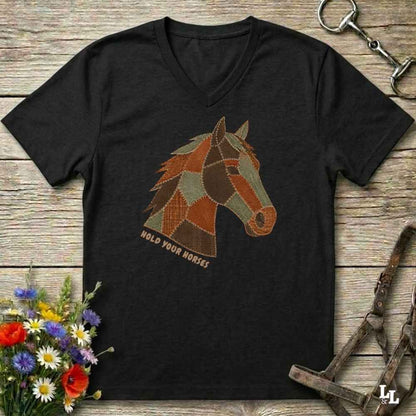 A black V-neck t-shirt laid flat on a rustic wooden surface, featuring a stylized patchwork horse head graphic with the text 'HOLD YOUR HORSES' underneath. The shirt is styled with equestrian tack, a small bouquet of wildflowers, and a metal horse bit.
