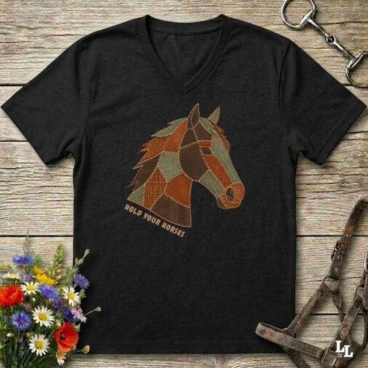 A black V-neck t-shirt laid flat on a rustic wooden surface, featuring a stylized patchwork horse head graphic with the text 'HOLD YOUR HORSES' underneath. The shirt is styled with equestrian tack, a small bouquet of wildflowers, and a metal horse bit.