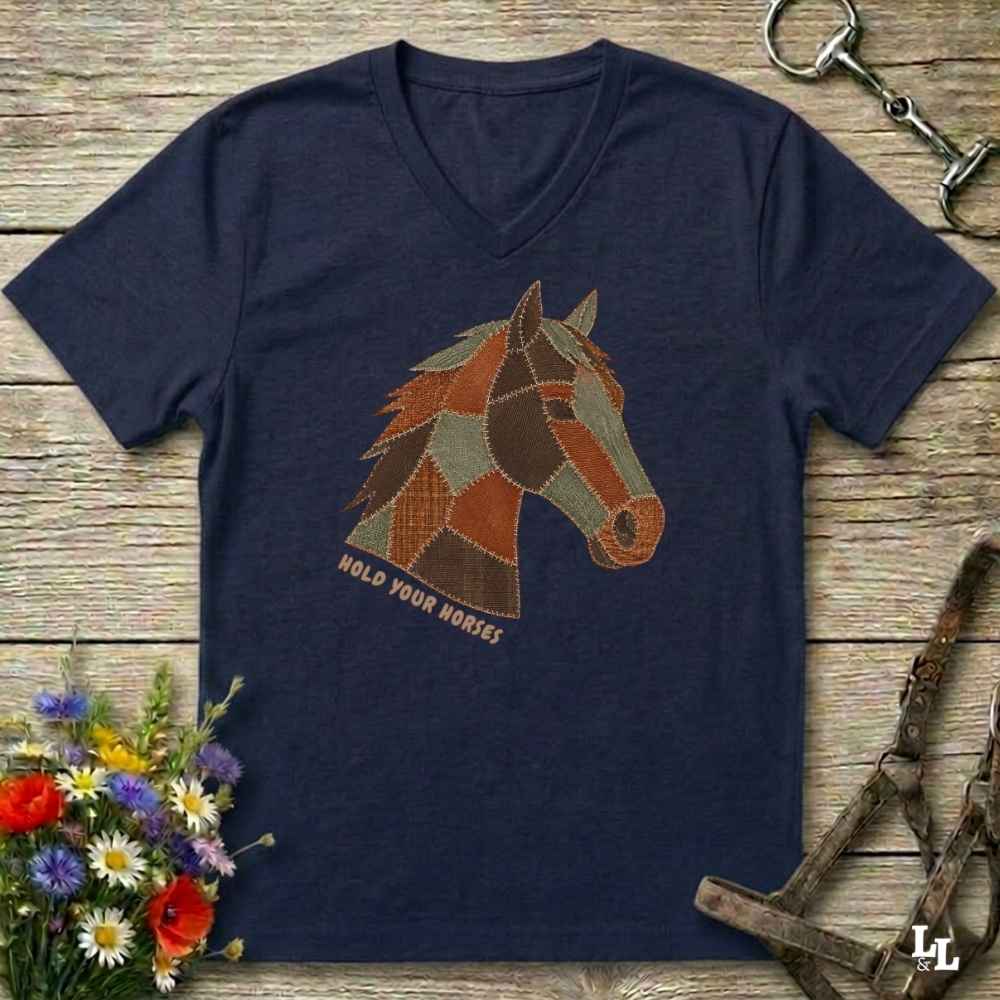 A navy blue V-neck t-shirt laid flat on a rustic wooden surface, featuring a stylized patchwork horse head graphic with the text 'HOLD YOUR HORSES' underneath. The shirt is styled with equestrian tack, a small bouquet of wildflowers, and a metal horse bit.