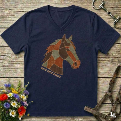 A navy blue V-neck t-shirt laid flat on a rustic wooden surface, featuring a stylized patchwork horse head graphic with the text 'HOLD YOUR HORSES' underneath. The shirt is styled with equestrian tack, a small bouquet of wildflowers, and a metal horse bit.