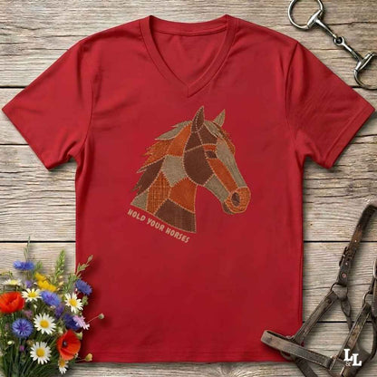 A red V-neck t-shirt laid flat on a rustic wooden surface, featuring a patchwork-style horse head graphic and the text 'HOLD YOUR HORSES' printed below it. The shirt is styled with equestrian-themed props including a horse bit, leather bridle, and a small bouquet of wildflowers.