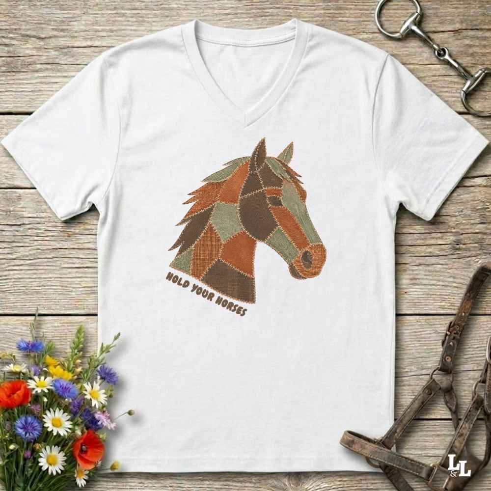 A white V-neck t-shirt laid flat on a rustic wooden surface, featuring a patchwork-style horse head graphic in brown, tan, and olive tones with the text 'HOLD YOUR HORSES' beneath it. The shirt is styled with equestrian-themed props including a halter, tack, and flowers.