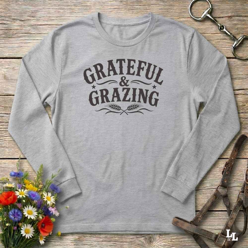 A light heather gray long-sleeve t-shirt with a dark brown graphic reading 'GRATEFUL & GRAZING' with decorative flourishes and wheat ears, laid flat on a rustic wooden surface with flowers and horse tack props.