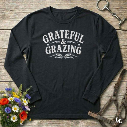 A black long-sleeve t-shirt with a white graphic print reading 'GRATEFUL & GRAZING' on the chest, styled in a flat-lay on a rustic wooden background with equestrian and floral props.