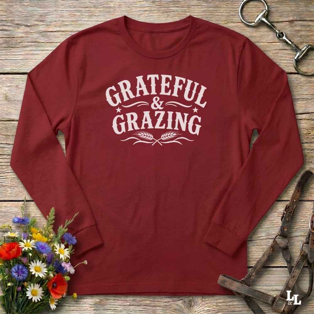 A red long-sleeve t-shirt with a dark brown graphic reading 'GRATEFUL & GRAZING' with decorative flourishes and wheat ears, laid flat on a rustic wooden surface with flowers and horse tack props.