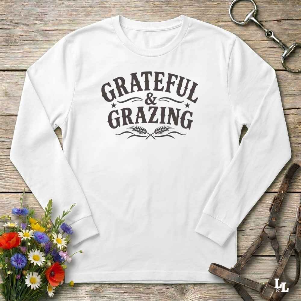 A white long-sleeve t-shirt with a dark brown graphic reading 'GRATEFUL & GRAZING' with decorative flourishes and wheat ears, laid flat on a rustic wooden surface with flowers and horse tack props.