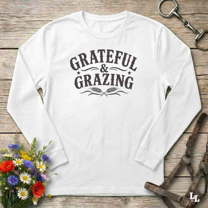A white long-sleeve t-shirt with a dark brown graphic reading 'GRATEFUL & GRAZING' with decorative flourishes and wheat ears, laid flat on a rustic wooden surface with flowers and horse tack props.
