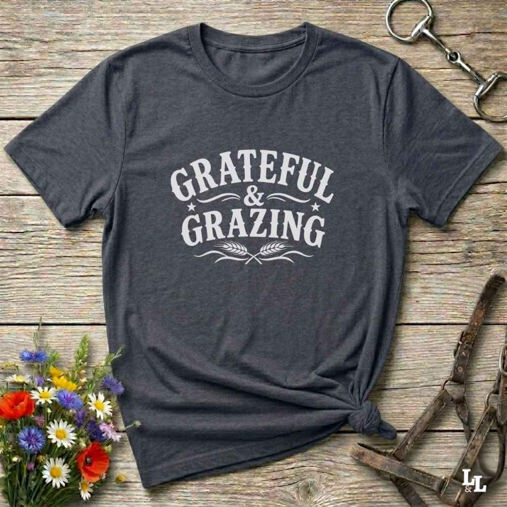 A dark heather gray t-shirt with a white graphic reading 'Grateful & Grazing' laid flat on a rustic wooden surface, styled with flowers, a horse bit, and a leather halter.