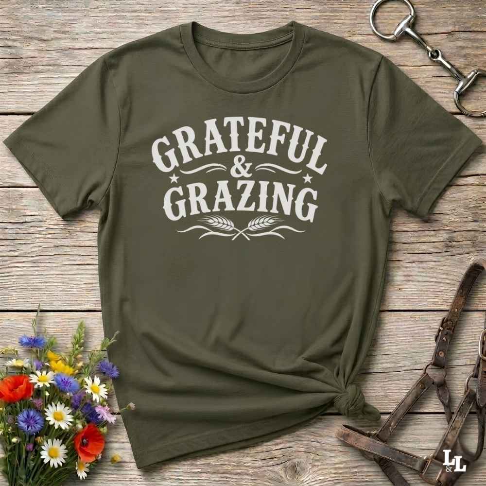 An olive green short-sleeve t-shirt with a white graphic print reading 'GRATEFUL & GRAZING' in a western-style font, styled in a flat-lay on a rustic wooden background with flowers and horse tack props.