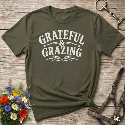 An olive green short-sleeve t-shirt with a white graphic print reading 'GRATEFUL & GRAZING' in a western-style font, styled in a flat-lay on a rustic wooden background with flowers and horse tack props.
