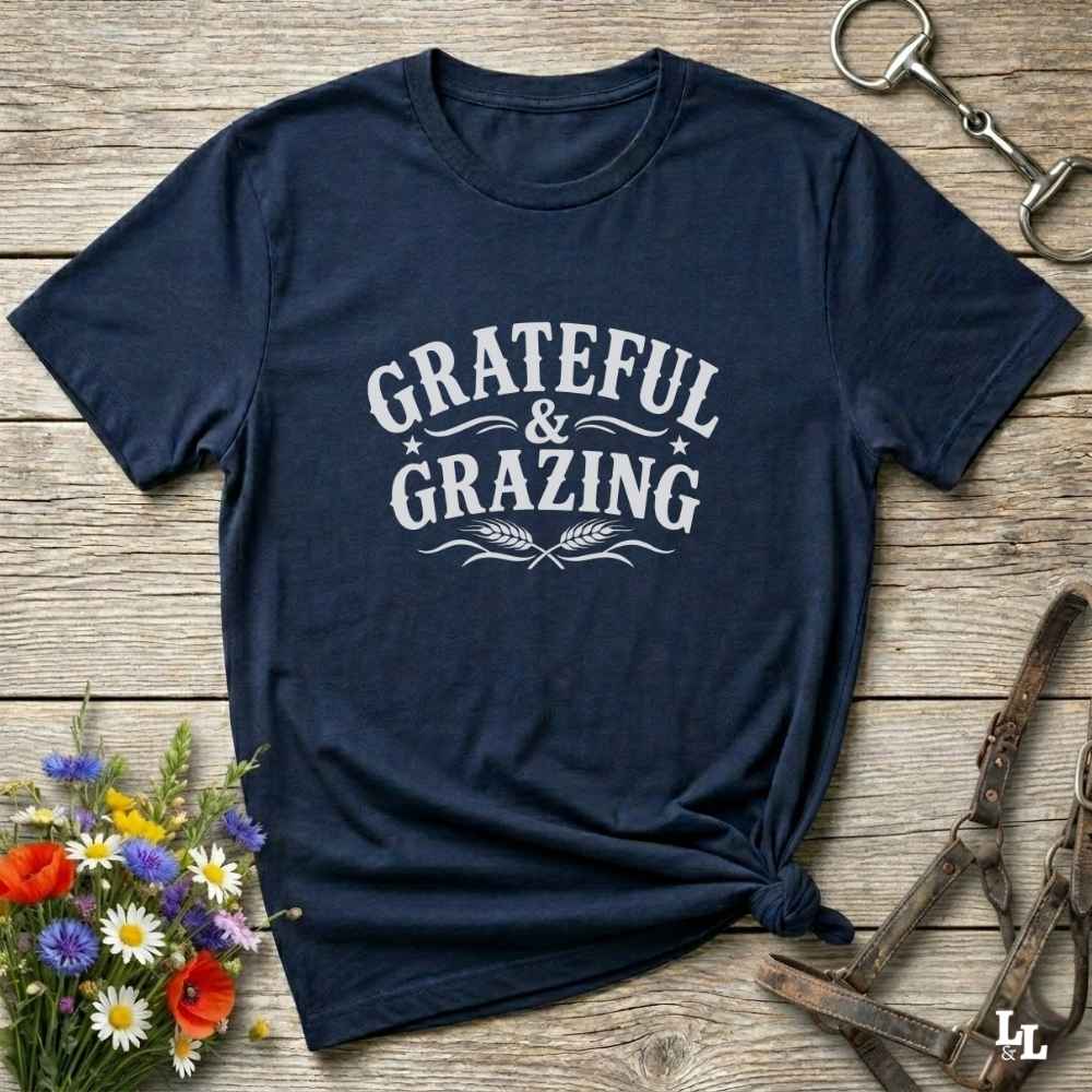 A navy blue short-sleeve t-shirt with a white graphic reading 'GRATEFUL & GRAZING' laid flat on a rustic wooden surface. The shirt is styled with a knot at the bottom hem, surrounded by decorative props including a small bouquet of wildflowers, a metal horse bit, and a leather halter.