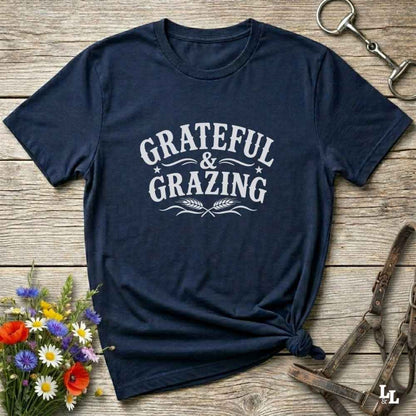 A navy blue short-sleeve t-shirt with a white graphic reading 'GRATEFUL & GRAZING' laid flat on a rustic wooden surface. The shirt is styled with a knot at the bottom hem, surrounded by decorative props including a small bouquet of wildflowers, a metal horse bit, and a leather halter.