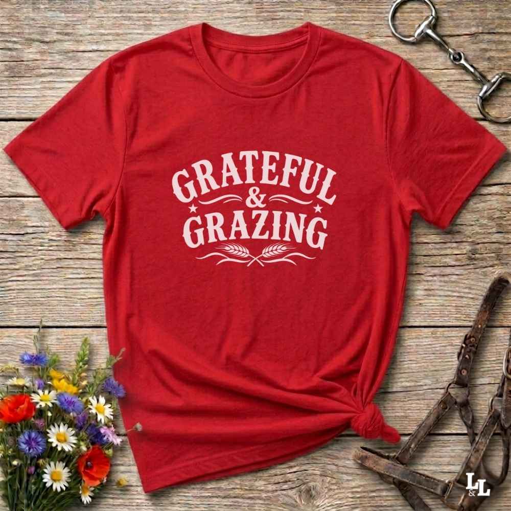 A red short-sleeve crewneck t-shirt with 'GRATEFUL & GRAZING' printed in white across the chest, styled flat on a rustic wooden surface with equestrian accessories and flowers.