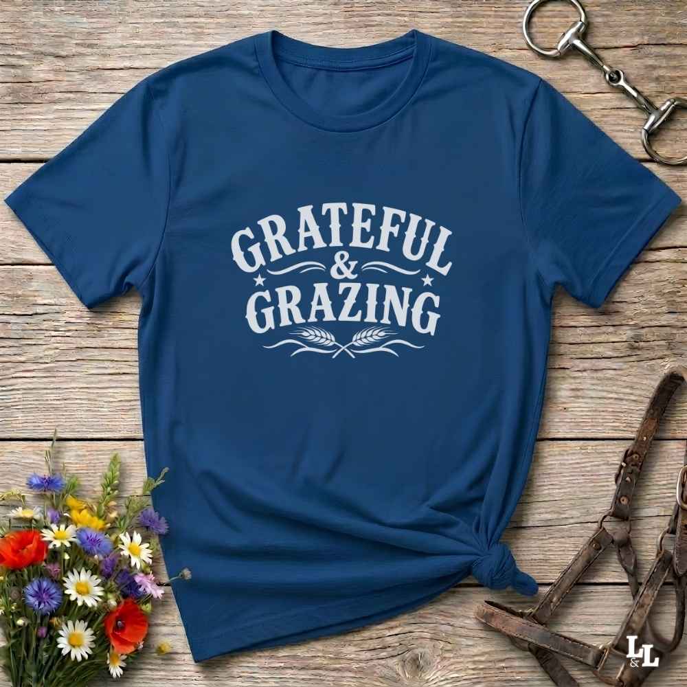 A navy blue short-sleeve t-shirt with a white graphic print reading 'GRATEFUL & GRAZING' and wheat illustrations, styled flat on a rustic wooden surface with flowers and horse tack props.