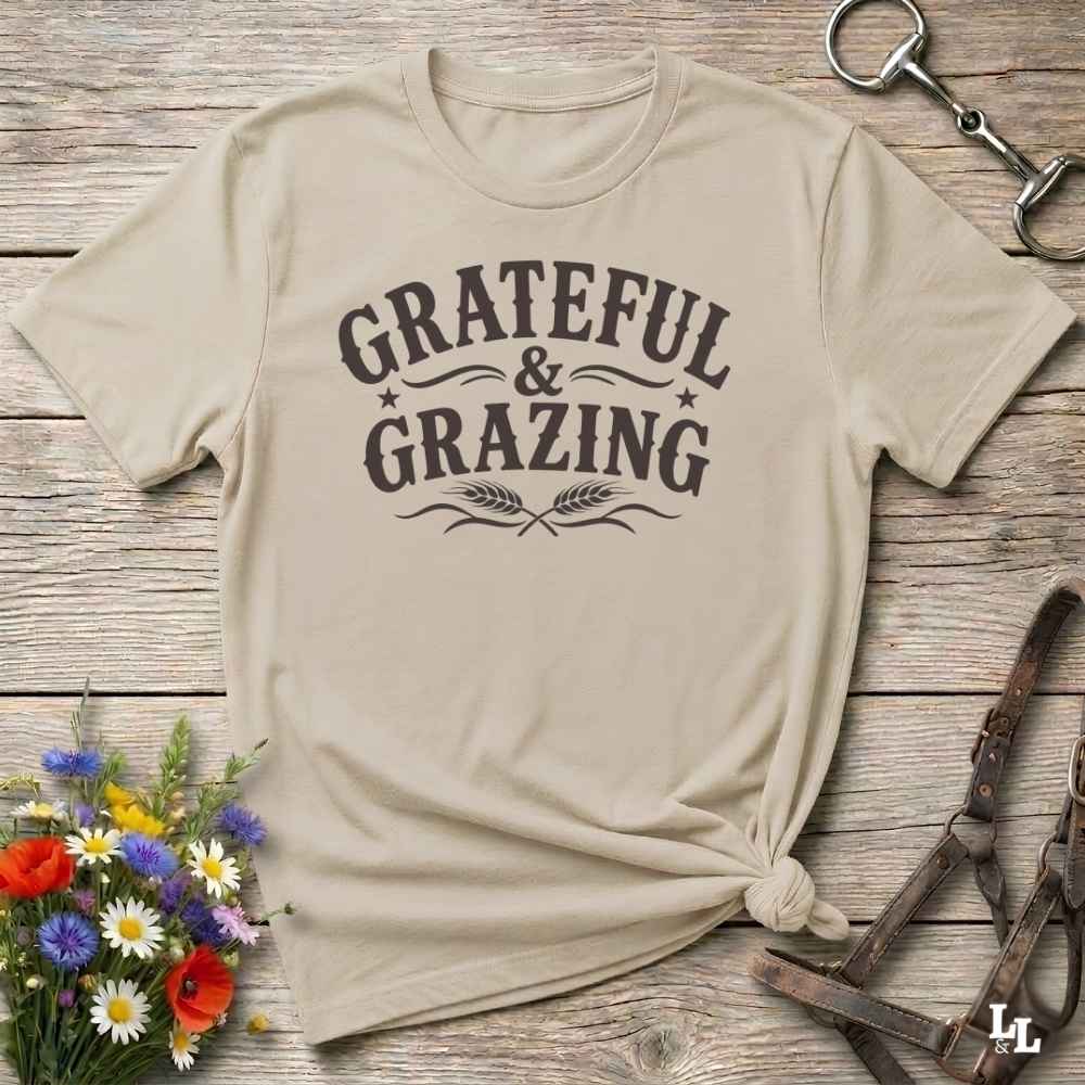 A beige short-sleeve t-shirt with a dark brown graphic reading 'GRATEFUL & GRAZING' laid flat on a rustic wooden surface, styled with flowers, a horse bit, and a leather strap.