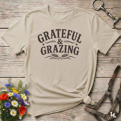 A beige short-sleeve t-shirt with a dark brown graphic reading 'GRATEFUL & GRAZING' laid flat on a rustic wooden surface, styled with flowers, a horse bit, and a leather strap.