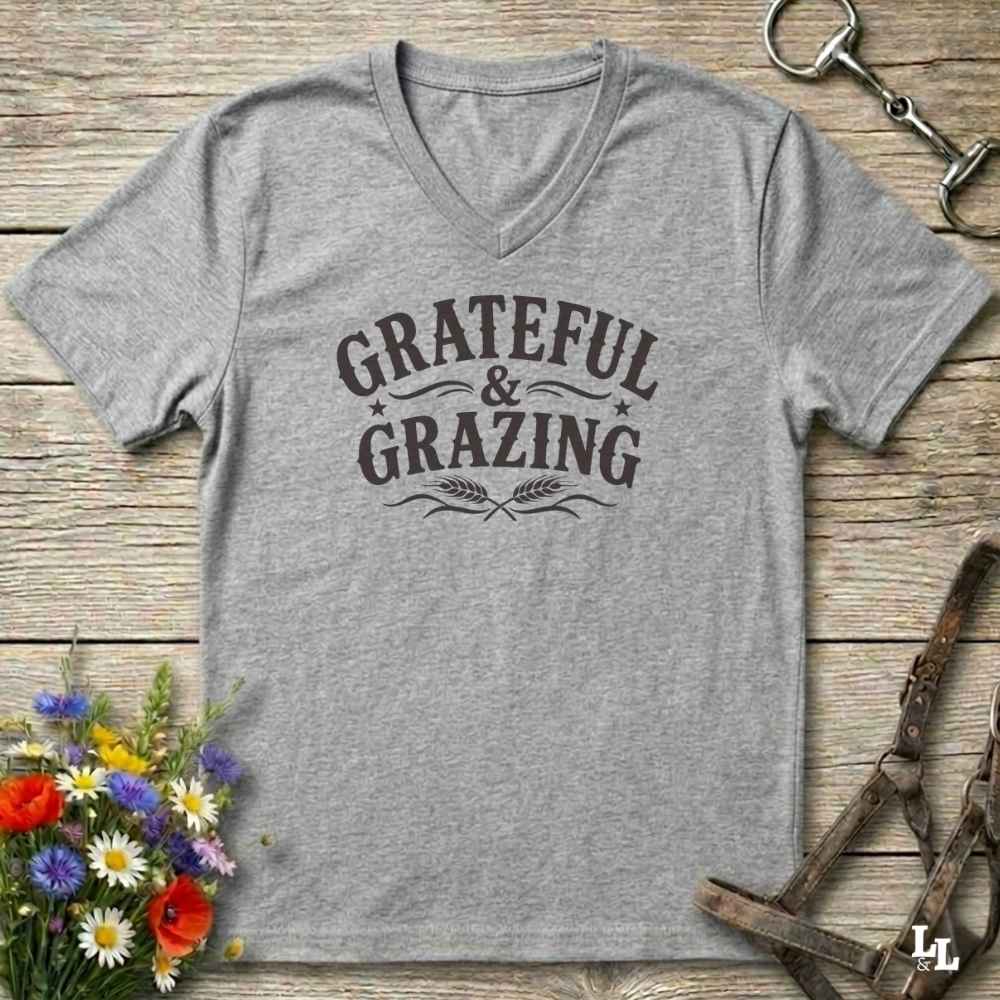 A heather gray V-neck t-shirt with a dark brown graphic reading 'GRATEFUL & GRAZING' with wheat stalks, laid flat on a rustic wooden surface with equestrian and floral props.