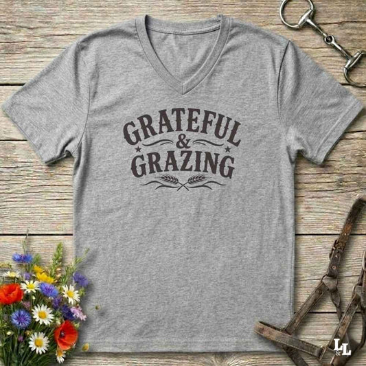A heather gray V-neck t-shirt with a dark brown graphic reading 'GRATEFUL & GRAZING' with wheat stalks, laid flat on a rustic wooden surface with equestrian and floral props.