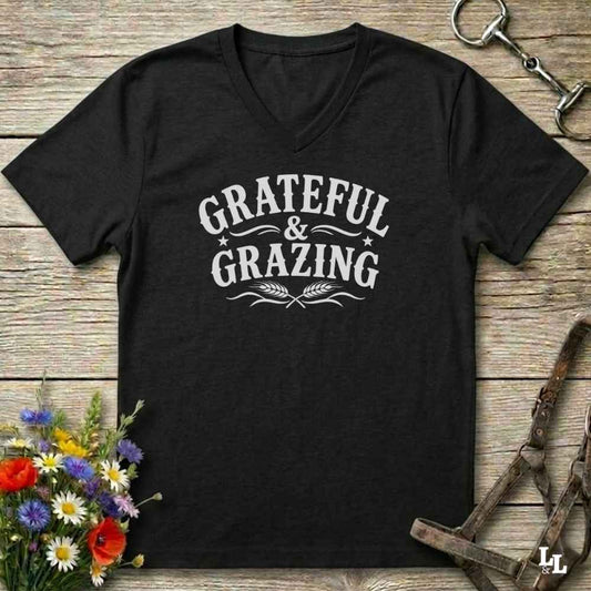 A black V-neck t-shirt with a white graphic reading 'GRATEFUL & GRAZING' and wheat stalks, laid flat on a rustic wood background with flowers and horse tack props.
