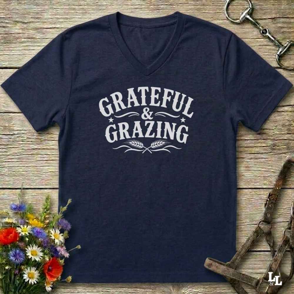 A navy blue V-neck t-shirt with a white graphic reading 'Grateful & Grazing' laid flat on a rustic wooden surface, styled with flowers, horse tack, and a bridle.