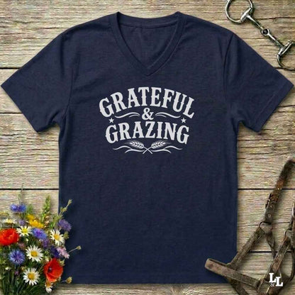 A navy blue V-neck t-shirt with a white graphic reading 'Grateful & Grazing' laid flat on a rustic wooden surface, styled with flowers, horse tack, and a bridle.