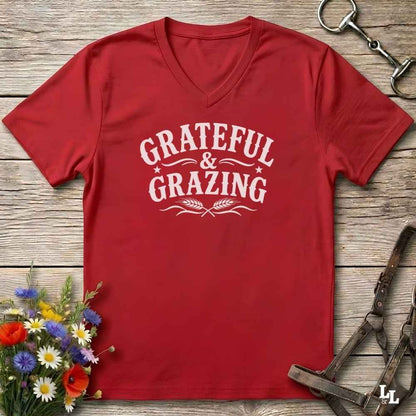 A red V-neck t-shirt with a white graphic reading 'Grateful & Grazing' laid flat on a rustic wooden surface, styled with flowers, horse tack, and a bridle.