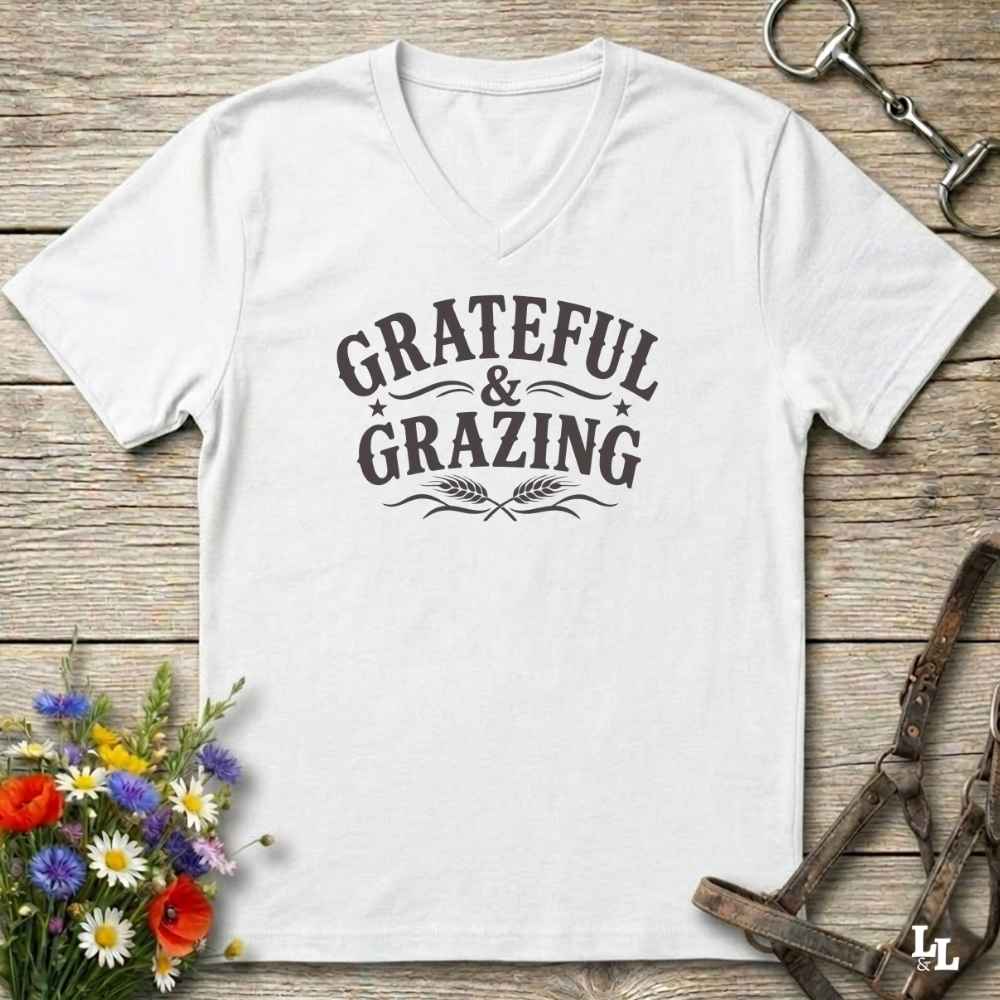 A white V-neck t-shirt with a dark gray graphic reading 'GRATEFUL & GRAZING' with decorative flourishes, laid flat on a rustic wooden surface with flowers and horse tack props.