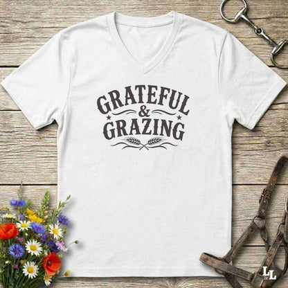 A white V-neck t-shirt with a dark gray graphic reading 'GRATEFUL & GRAZING' with decorative flourishes, laid flat on a rustic wooden surface with flowers and horse tack props.