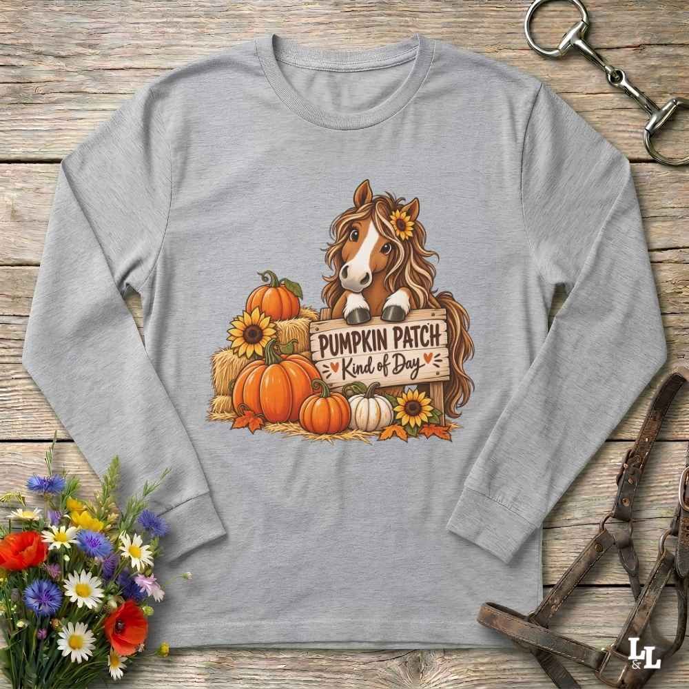 A heather gray long-sleeve t-shirt laid flat on a rustic wooden surface, featuring a graphic of a brown horse with a sunflower in its mane, surrounded by pumpkins, sunflowers, and a sign that reads 'PUMPKIN PATCH = Kind of Dog'. Decorative flowers and horse tack are placed around the shirt.