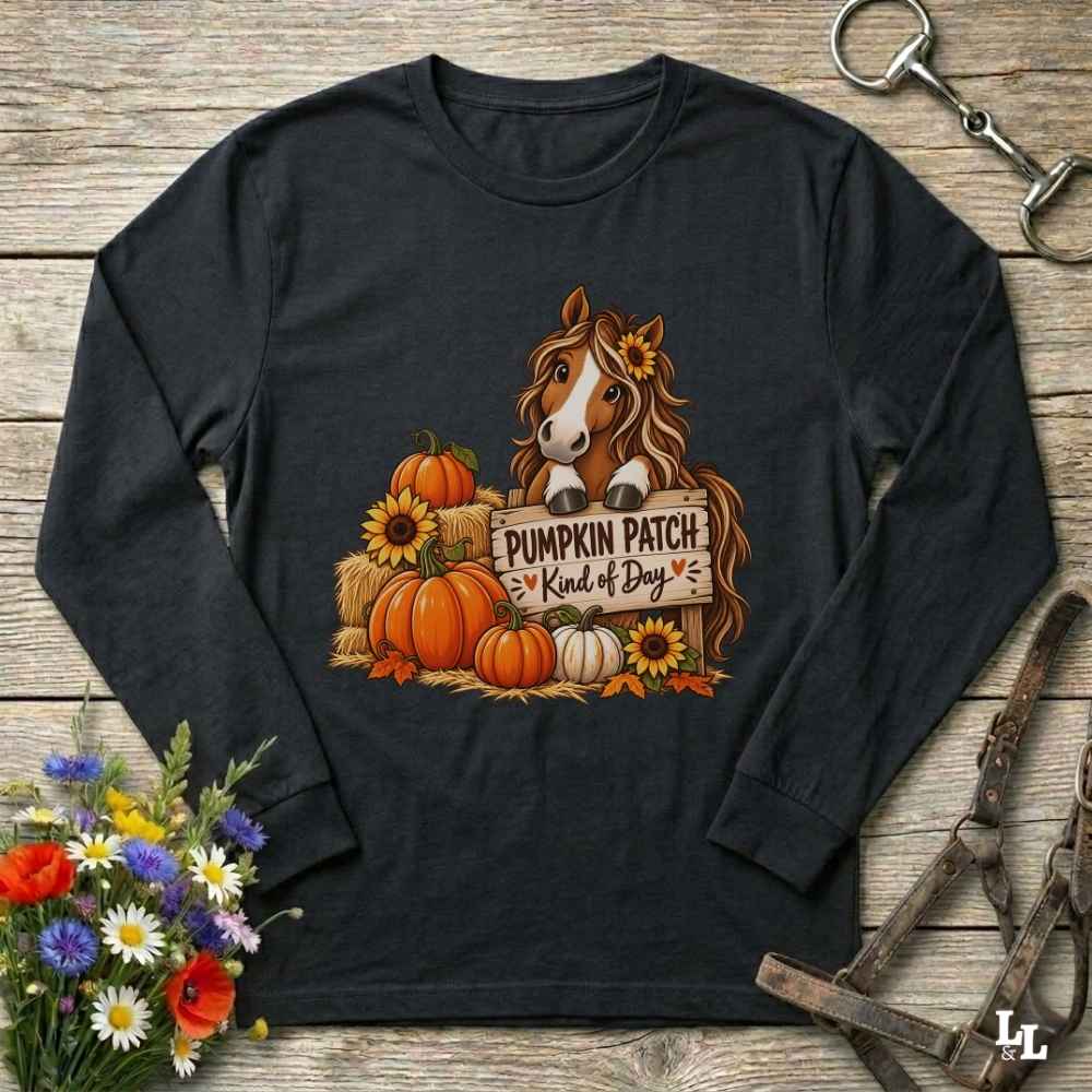A black long-sleeve t-shirt with a autumn-themed graphic of a horse behind pumpkins and sunflowers holding a sign that reads 'PUMPKIN PATCH Kind of Day'. The shirt is displayed flat on a rustic wooden background with decorative flowers and horse tack.