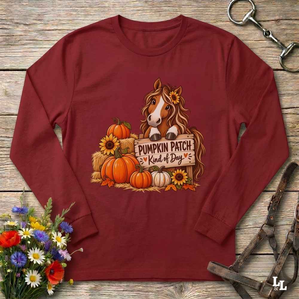 A long-sleeve crewneck t-shirt in maroon red, laid flat on a rustic wooden background. The shirt features a graphic of a brown horse behind a wooden sign that reads 'PUMPKIN PATCH = Kind of Dog', surrounded by pumpkins, sunflowers, and hay.