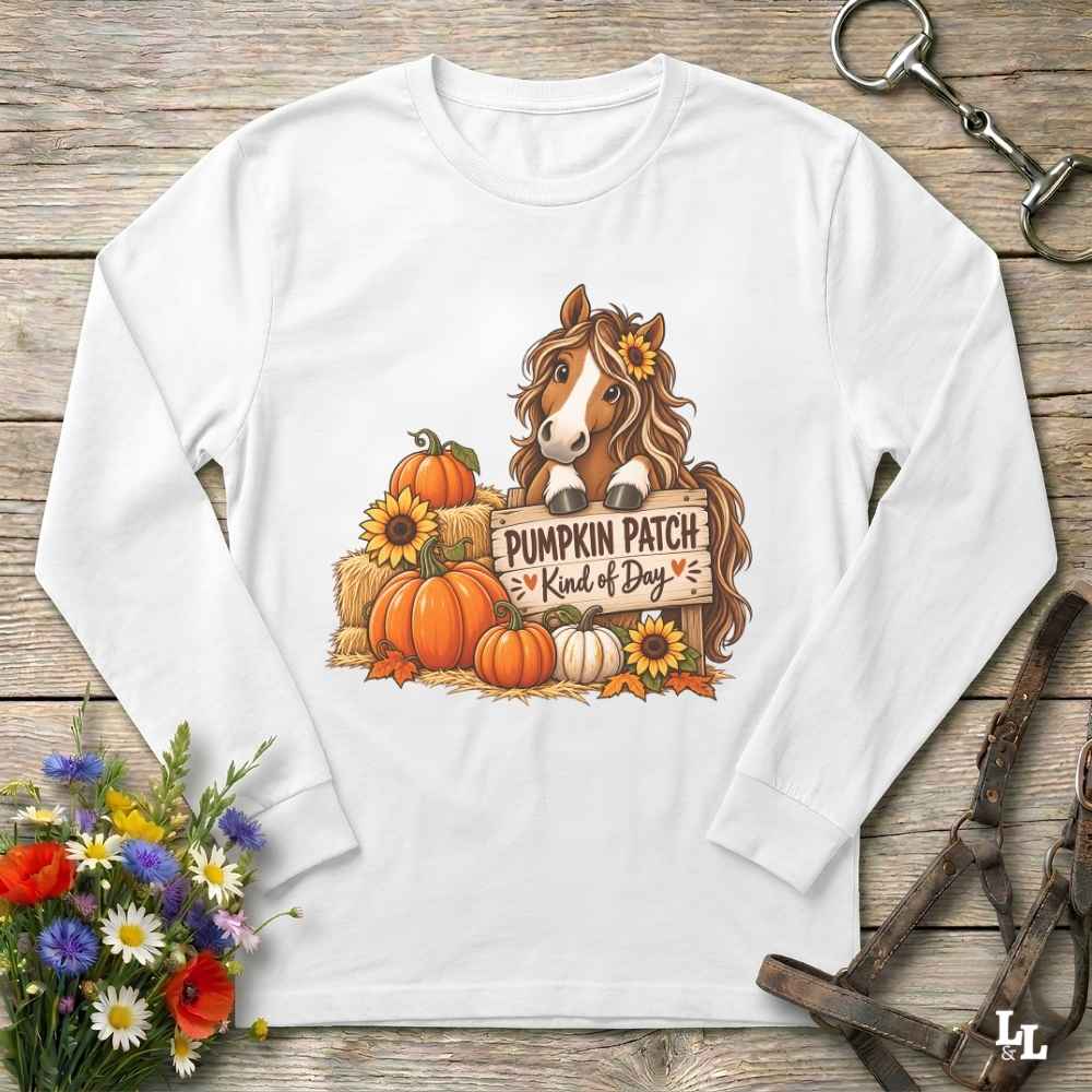 A white long-sleeve t-shirt with a autumn-themed graphic of a horse behind pumpkins and sunflowers holding a sign that reads 'PUMPKIN PATCH Kind of Day'. The shirt is displayed flat on a rustic wooden background with decorative flowers and horse tack.