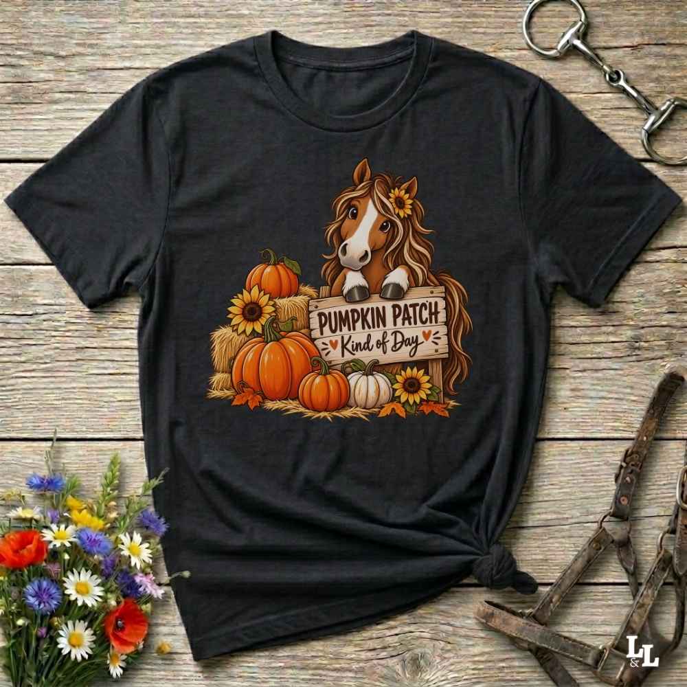 A black graphic t-shirt laid flat on a rustic wooden surface, styled with equestrian tack and a small bouquet of wildflowers. The shirt features a graphic of a brown horse with a sunflower in its hair, standing behind a sign that reads 'PUMPKIN PATCH Kind of Day' with orange pumpkins and yellow sunflowers.