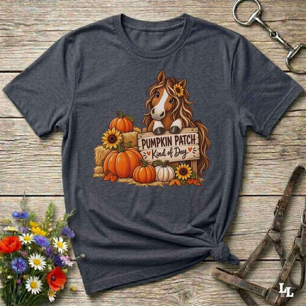 A dark heather gray graphic t-shirt laid flat on a rustic wooden surface, styled with equestrian tack and a small bouquet of wildflowers. The graphic features a brown horse with a sunflower in its hair, standing behind a sign that reads 'PUMPKIN PATCH is Kind of Day' with orange, white, and yellow pumpkins and sunflowers.