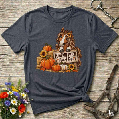 A dark heather gray graphic t-shirt laid flat on a rustic wooden surface, styled with equestrian tack and a small bouquet of wildflowers. The graphic features a brown horse with a sunflower in its hair, standing behind a sign that reads 'PUMPKIN PATCH is Kind of Day' with orange, white, and yellow pumpkins and sunflowers.
