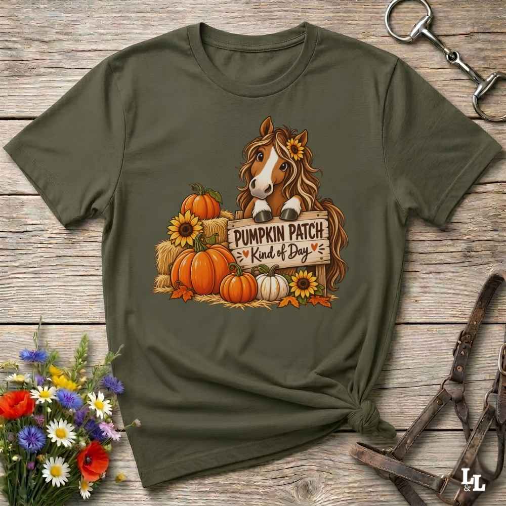 An olive green short-sleeve t-shirt with a autumn-themed graphic of a brown horse behind pumpkins and sunflowers with the text 'PUMPKIN PATCH is Kind of Day'. The shirt is styled in a flat-lay on a rustic wooden background with decorative flowers and leather tack.