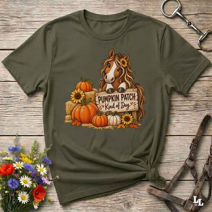 An olive green short-sleeve t-shirt with a autumn-themed graphic of a brown horse behind pumpkins and sunflowers with the text 'PUMPKIN PATCH is Kind of Day'. The shirt is styled in a flat-lay on a rustic wooden background with decorative flowers and leather tack.