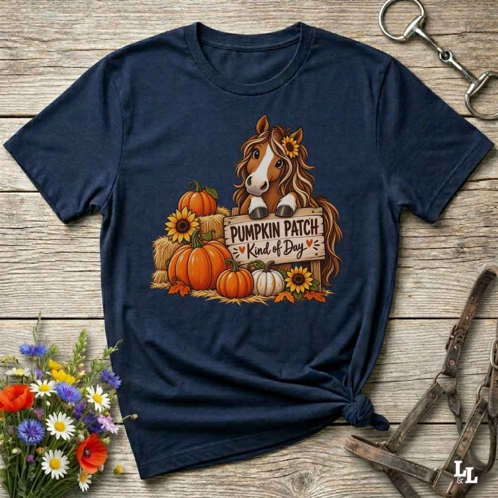 A navy blue graphic t-shirt laid flat on a rustic wooden surface, styled with equestrian tack and a small bouquet of wildflowers. The shirt features a graphic of a brown horse with a sunflower in its hair, standing behind a sign that reads 'PUMPKIN PATCH Kind of Day' with orange pumpkins and yellow sunflowers.