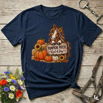 A navy blue graphic t-shirt laid flat on a rustic wooden surface, styled with equestrian tack and a small bouquet of wildflowers. The shirt features a graphic of a brown horse with a sunflower in its hair, standing behind a sign that reads 'PUMPKIN PATCH Kind of Day' with orange pumpkins and yellow sunflowers.