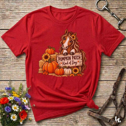 A red graphic t-shirt laid flat on a rustic wooden surface, styled with equestrian tack and a small bouquet of wildflowers. The shirt features a graphic of a brown horse with a sunflower in its hair, standing behind a sign that reads 'PUMPKIN PATCH Kind of Day' with orange pumpkins and yellow sunflowers.