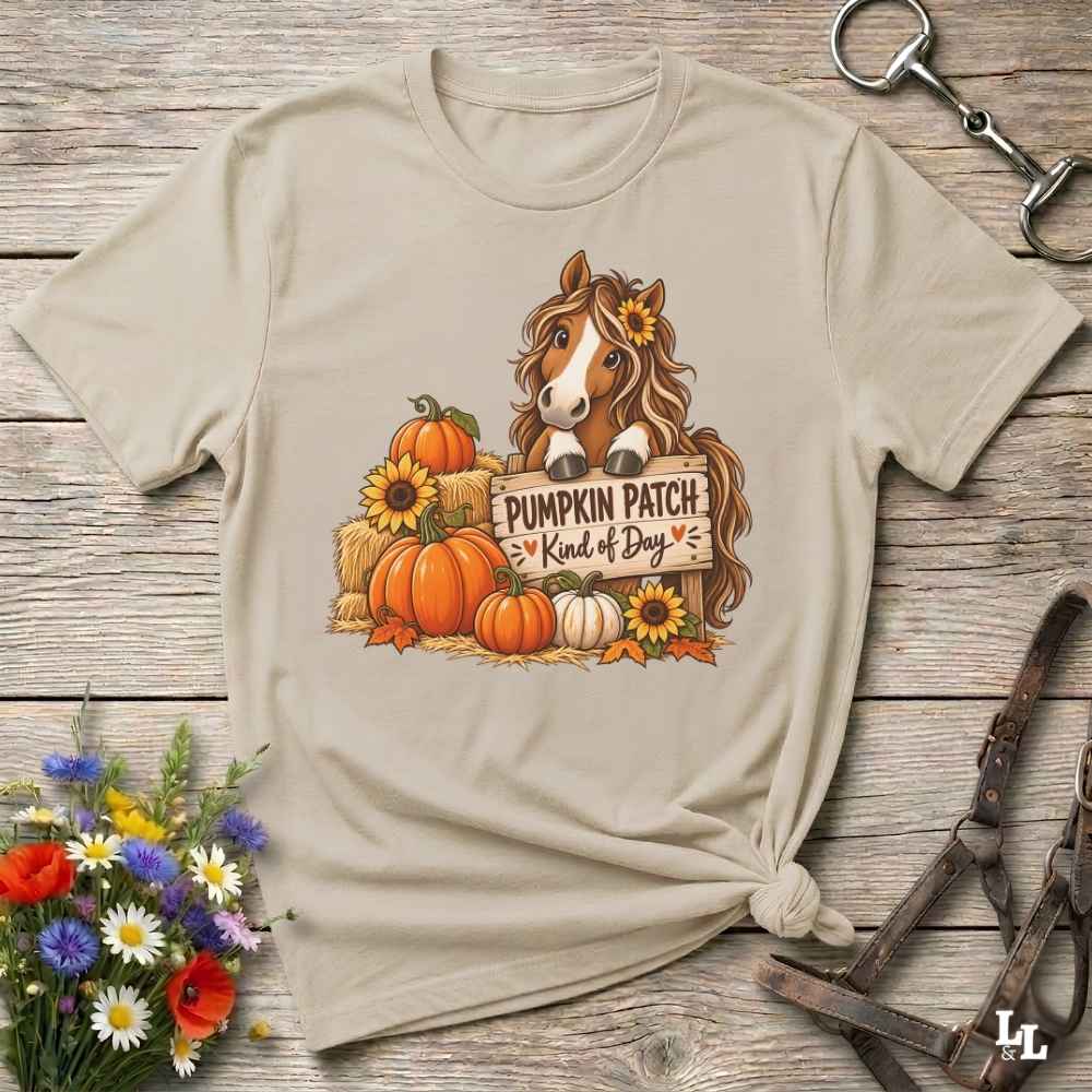 A beige graphic t-shirt featuring a cartoon horse surrounded by pumpkins, sunflowers, and a sign that reads 'PUMPKIN PATCH Kind of Day'. The shirt is styled in a flat-lay on a rustic wooden background with horse tack and flowers.