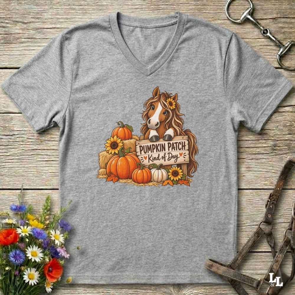 A greV-neck t-shirt laid flat on a rustic wooden surface, featuring a graphic of a brown horse behind a sign that reads 'PUMPKIN PATCH Kind of Boy' surrounded by pumpkins, sunflowers, and hay bales. The shirt is styled with horse tack and flowers around it. 