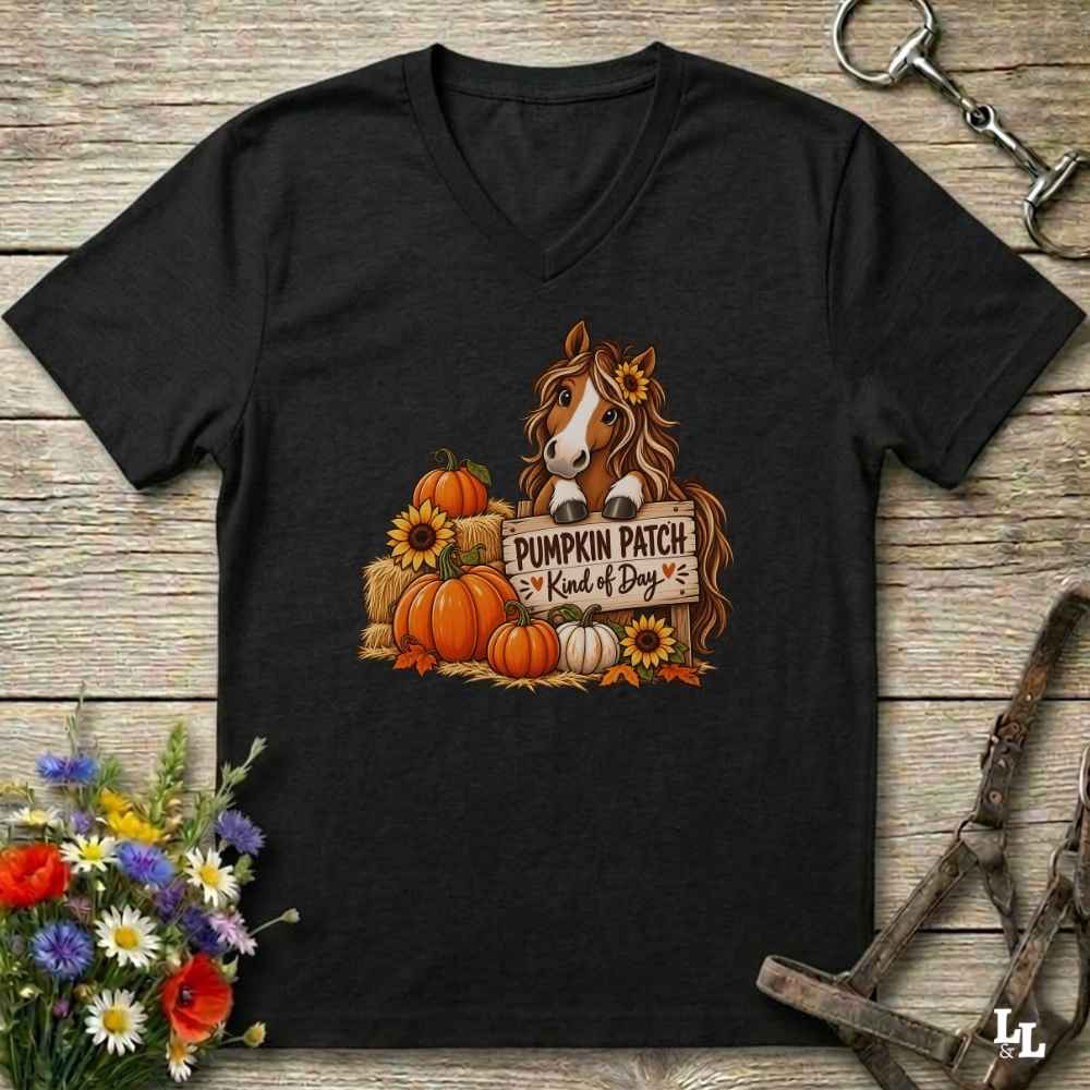 A black V-neck t-shirt laid flat on a rustic wooden surface, featuring a graphic of a brown horse behind a sign that reads 'PUMPKIN PATCH Kind of Boy' surrounded by pumpkins, sunflowers, and hay bales. The shirt is styled with horse tack and flowers around it.