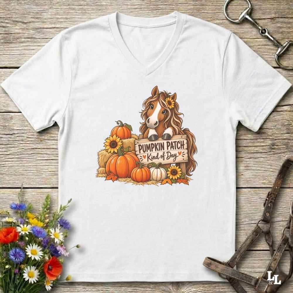 A white V-neck t-shirt laid flat on a rustic wooden surface, featuring a colorful graphic of a horse with a flower in its hair standing among pumpkins and sunflowers next to a sign that reads 'Pumpkin Patch Kind of Dog'. Decorative flowers and horse tack are placed around the shirt.