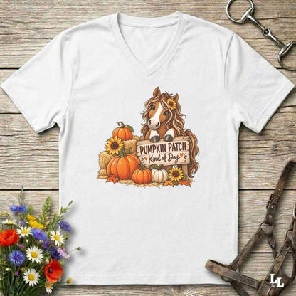 A white V-neck t-shirt laid flat on a rustic wooden surface, featuring a colorful graphic of a horse with a flower in its hair standing among pumpkins and sunflowers next to a sign that reads 'Pumpkin Patch Kind of Dog'. Decorative flowers and horse tack are placed around the shirt.
