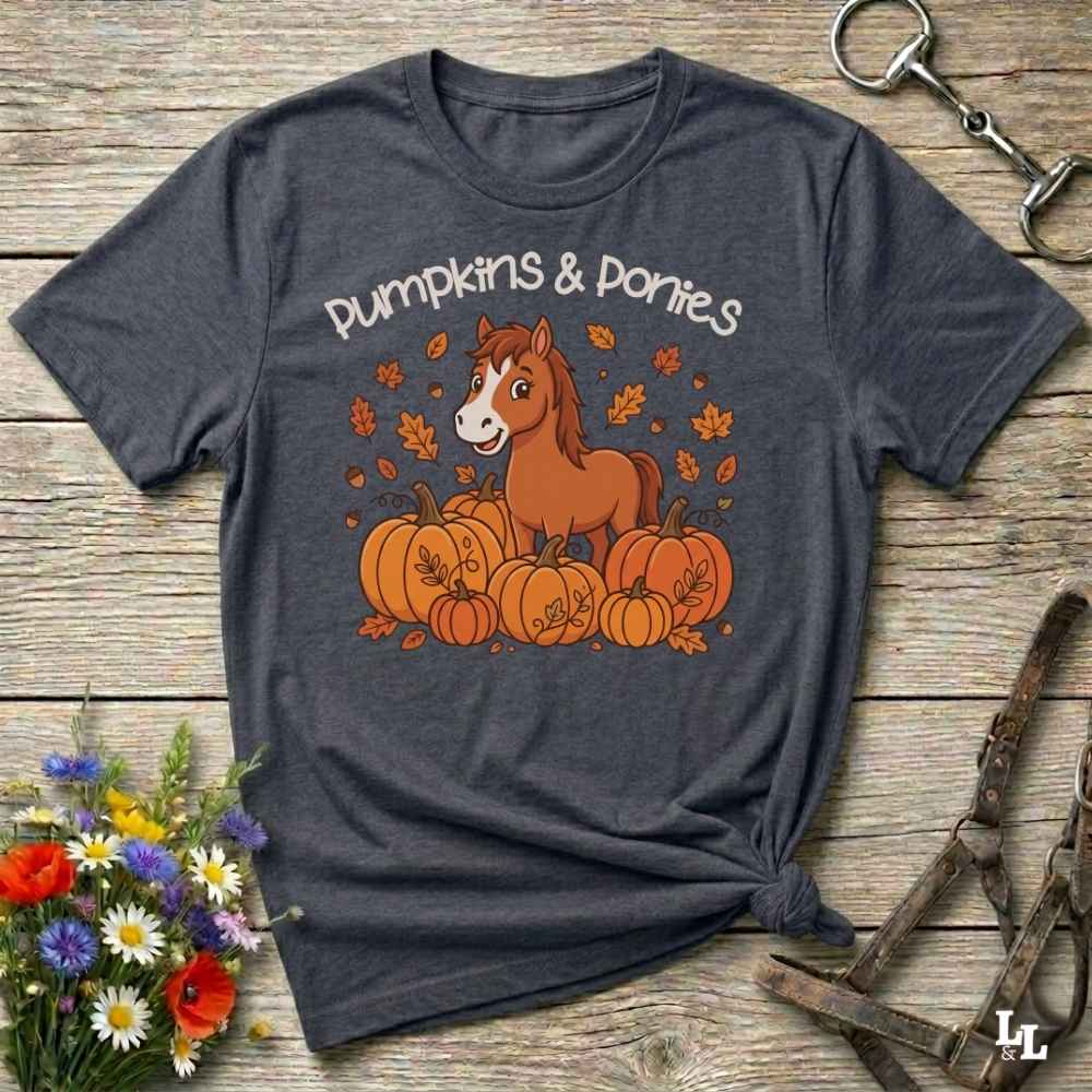 A dark heather gray graphic t-shirt laid flat on a rustic wooden surface, styled with autumn props including a bouquet of wildflowers, a horse halter, and a metal ring. The t-shirt features a cartoon illustration of a brown horse standing among orange pumpkins and falling leaves, with the text 'Pumpkins & Ponies' written above.