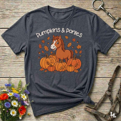A dark heather gray graphic t-shirt laid flat on a rustic wooden surface, styled with autumn props including a bouquet of wildflowers, a horse halter, and a metal ring. The t-shirt features a cartoon illustration of a brown horse standing among orange pumpkins and falling leaves, with the text 'Pumpkins & Ponies' written above.