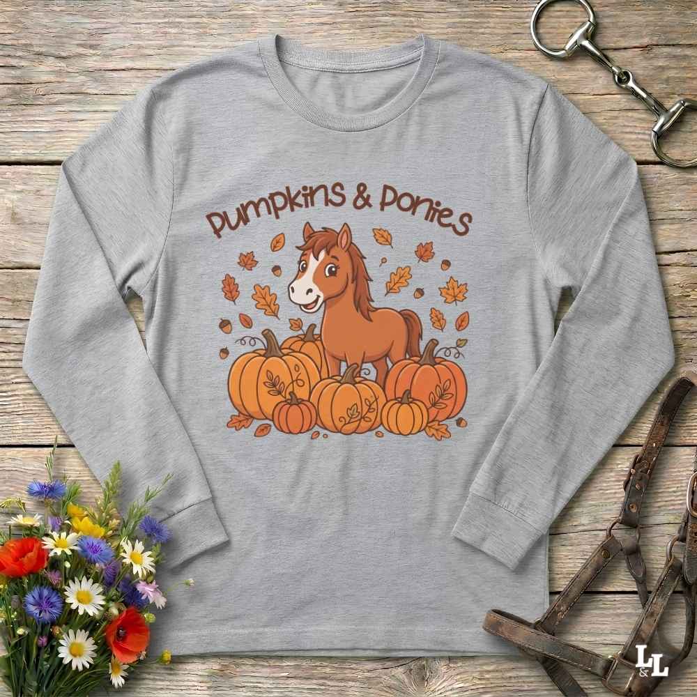 A grey long-sleeve t-shirt with a 'Pumpkins & Ponies' graphic, laid flat on a rustic wooden background with decorative autumn props including flowers, a horse bit, and a leather bridle.
